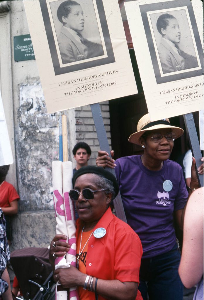 Two Black Lesbians among a group holding signs that read "Lesbian Herstory Archives: In Memory of the Voices We Have Lost."