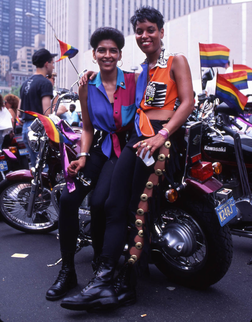 Two Black dykes sitting on a motorcycle at a march with people waving rainbow flags in the background.