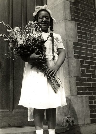 Audre Lorde as a young girl in a communion dress, holding a bouquet of flowers and standing on the steps in front of a door.