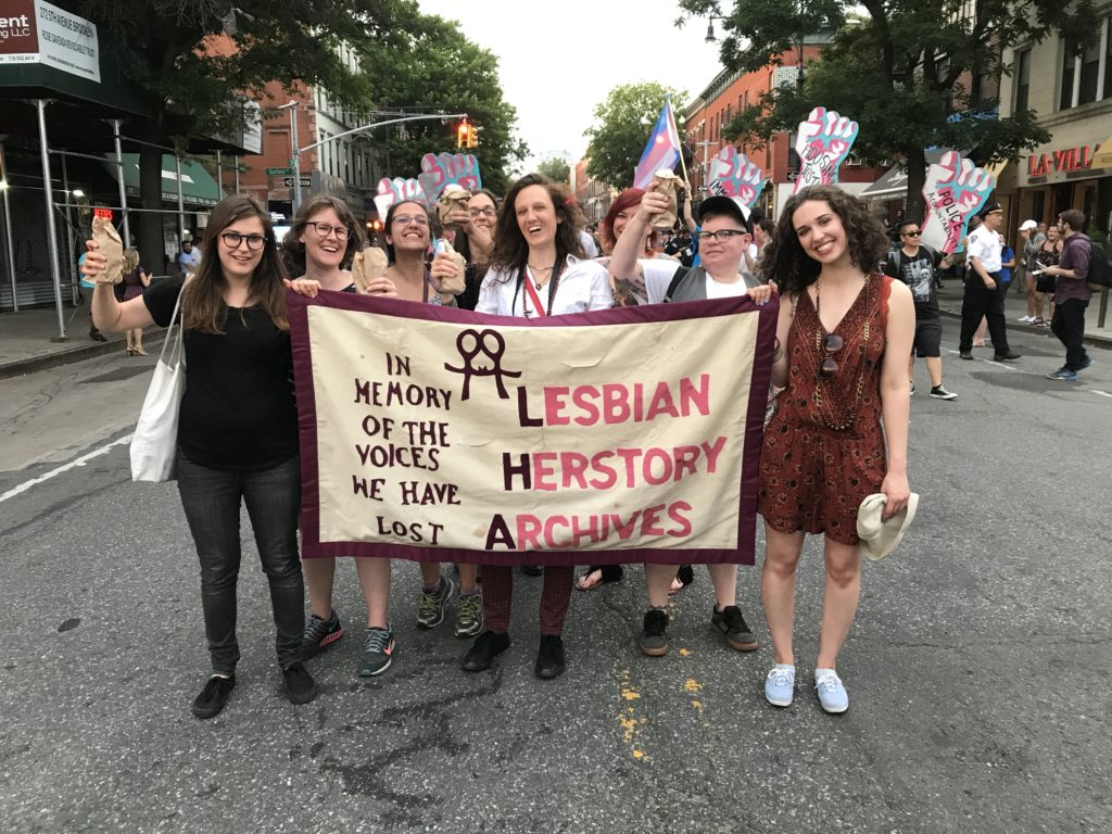 A group of Lesbians at a march holding a banner that reads "Lesbian Herstory Archives: In Memory of the Voices We Have Lost."