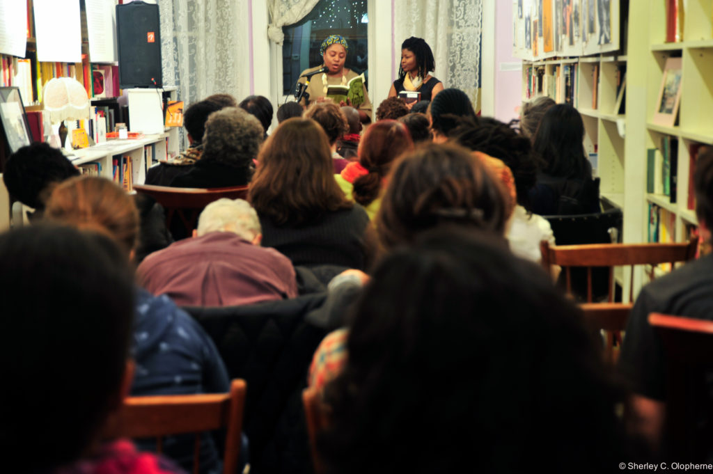 Two black lesbian activists at the front of the room holding a book affront a microphone facing a packed audience of women. Photo is taken from the back of the heads of the audience.